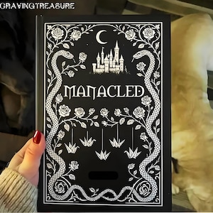 May include: Black book cover with white floral and snake illustrations, the word "MANACLED" in a decorative font, and origami cranes. A castle and crescent moon are also featured in the design. The book is held by a person.