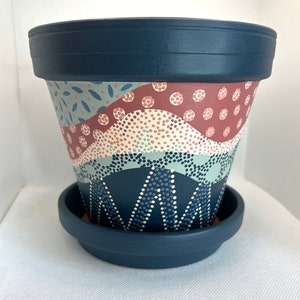 Op de afbeelding: Een handbeschilderde terracotta plantenpot met een geometrisch patroon in blauw, wit en roze. De pot heeft een bijpassende schotel.