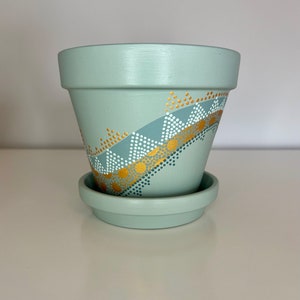May include: A light green terracotta planter with a gold and white geometric design. The planter has a matching saucer.
