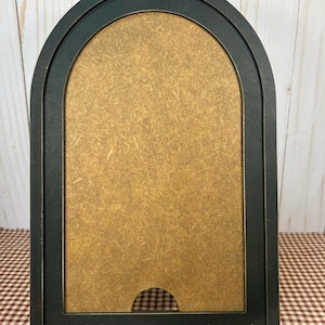 May include: An arched wooden frame with a matte black finish and a light brown fiberboard center panel. The frame features a small semi-circular cutout at the bottom edge. The text Muddygirlwoodcrafts is printed in white at the bottom center.