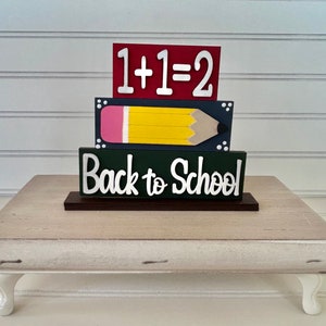 May include: A wooden tiered sign with the words "Back to School" in white on a green background, a yellow pencil with a pink eraser on a blue background, and the equation "1+1=2" in white on a red background.