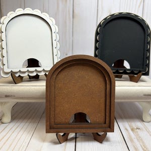 May include: Three small, decorative wooden frames in white, black, and brown. Each frame has an arched top and a scalloped edge. The frames are displayed on a white, tiered stand with curved legs. The frames are designed to stand upright.