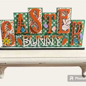 May include: Wooden block sign with the words "Easter Bunny" in orange letters on a green and white checkered background. The sign is sitting on a white wooden shelf with decorative legs.