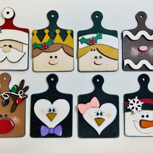 May include: Eight wooden cutting boards decorated with Christmas characters. The characters include Santa Claus, a gingerbread person, a reindeer, a snowman, and two penguins. The boards are painted in various colors, including red, green, white, brown, and black.