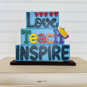 May include: A wooden sign with the words "Love Teach Inspire" in colorful letters. The sign is decorated with glitter and red hearts.