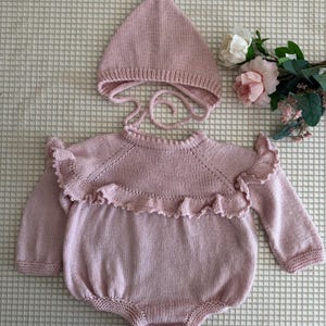 Könnte beinhalten: Ein hellrosa gestrickter Baby-Strampler mit langen Ärmeln und eine passende Mütze. Der Strampler hat einen gerüschten Besatz über der Brust und zwei kleine Knöpfe am unteren Ende. Die Mütze hat Bindebänder. Das Set ist aus weichem Strickstoff.
