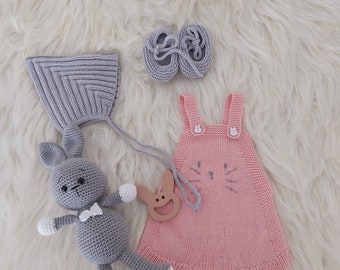 May include: A pink knitted baby onesie with a cat face design, a grey knitted hat with a matching bunny toy, and a pair of grey knitted booties.