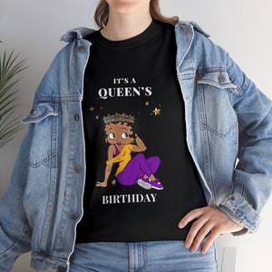 Puede incluir: Camiseta negra con una imagen de dibujos animados de una mujer con una corona y ropa morada y amarilla. El texto "It's a Queen's Birthday" está impreso en la camiseta.