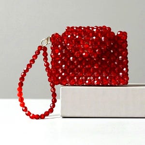 Può includere: Una pochette rettangolare rossa con perline e un cinturino da polso abbinato. La pochette è realizzata con piccole perline rosse sfaccettate e il cinturino è fissato con una chiusura color oro. La borsa è esposta su una superficie bianca su sfondo grigio.