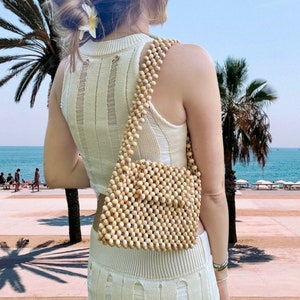 May include: A rectangular handbag constructed entirely from small, light-colored wooden beads, featuring a matching beaded shoulder strap. The bag has a uniform grid pattern created by the interconnected beads, providing a textured, natural appearance.