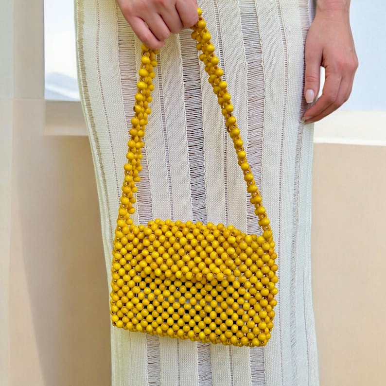 May include: A rectangular handbag constructed entirely from small, round, mustard yellow beads. The bag features a matching beaded shoulder strap and a structured, grid-like pattern created by the interconnected beads throughout the entire surface.