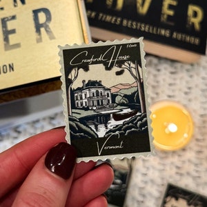 May include: A close-up of a book titled "Verity" by Colleen Hoover. The book is a collector's edition and features a gold cover with a black and white image of a woman. The book is sitting on a white surface with a small, white, wooden bead garland and a vintage-style stamp with a picture of a house and the words "Goddard House" and "Vermont".