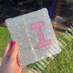 Könnte beinhalten: Ein kleines, silbernes, glitzerndes Notizbuch mit einem rosafarbenen Buchstaben "I" auf der Vorderseite. Das Notizbuch ist mit Strasssteinen besetzt.