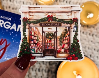 Puede incluir: Una pegatina de una librería con una corona de Navidad sobre la puerta. La librería está decorada con luces de Navidad y adornos. La pegatina está sobre una superficie blanca y peluda con velas encendidas.