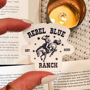 May include: A close-up of a white sticker with black text and a black and white illustration of a cowboy riding a horse. The sticker says "Rebel Blue Ranch" and "Est. 2023".