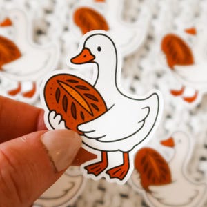 May include: A collection of stickers featuring a white goose with an orange beak and feet, holding a loaf of bread. The stickers have a white border and are on a textured white background.