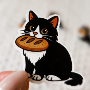 May include: A sticker featuring a black and white cat with orange ear tips, holding a round loaf of bread in its mouth. The cat has a white patch on its chest and is sitting upright. The sticker is on a white background.