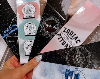 May include: A set of six colorful bookmarks featuring different designs. The bookmarks are made of cardstock and have a glossy finish. The designs include a zodiac sign, a book club logo, a constellation, and a series of globes with the words "Aer", "Ignis", "Aqua", and "Terra".
