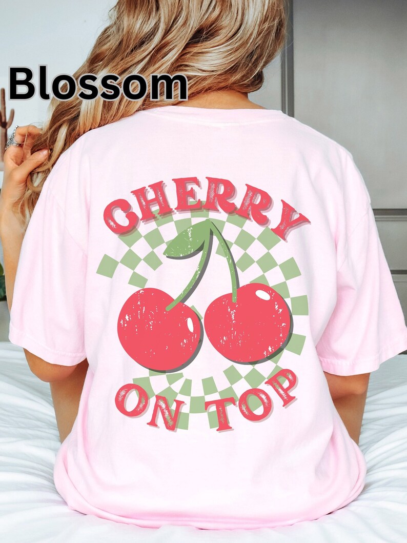 May include: Pink t-shirt with a green and white checkered design and a red cherry graphic. The text "CHERRY ON TOP" is printed on the shirt.