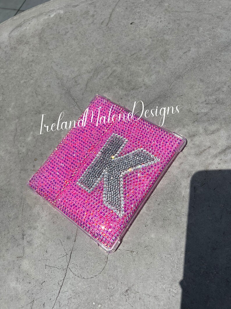 May include: A pink phone case with a clear back and a silver glitter letter K on the front. The case is covered in small pink glitter. The text "Ireland Malone Designs" is visible on the case.