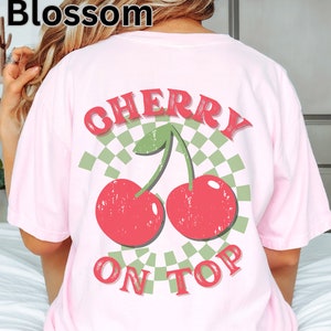 May include: Pink t-shirt with a graphic of two red cherries with green stems and leaves on a green and white checkered background. The text "CHERRY ON TOP" is printed below the cherries.