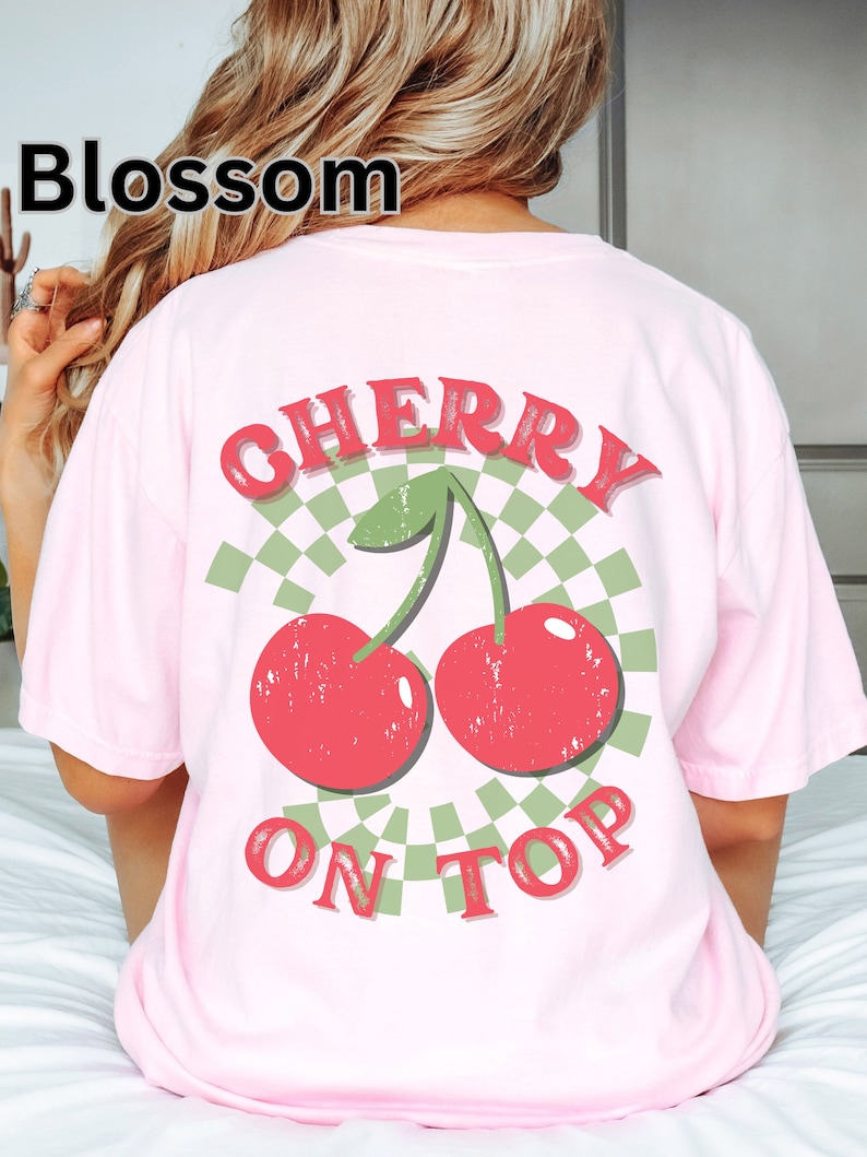 May include: Pink t-shirt with a graphic of two red cherries with green stems and leaves on a green and white checkered background. The text "CHERRY ON TOP" is printed below the cherries.
