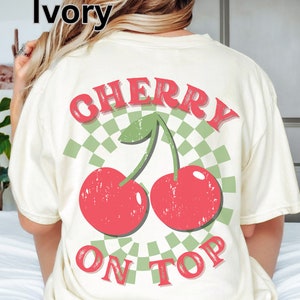 May include: Ivory t-shirt with a graphic of two red cherries with green stems and leaves on a green and white checkered background. The text "CHERRY ON TOP" is printed in red on the shirt.
