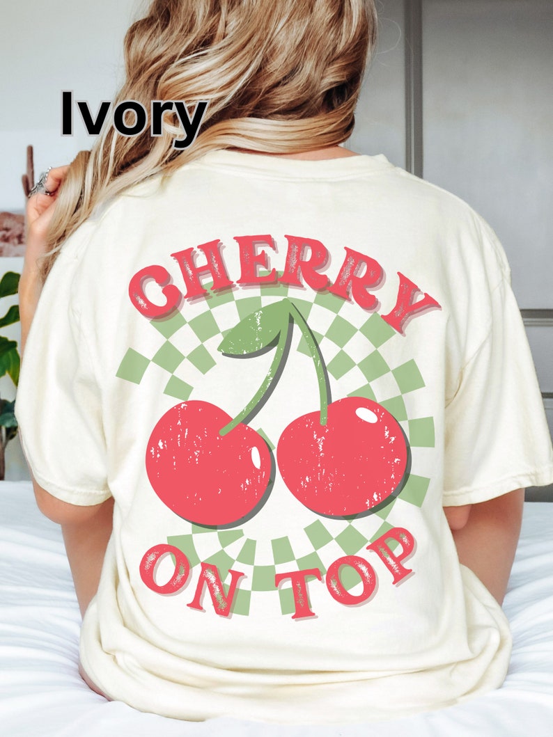 May include: Ivory t-shirt with a graphic of two red cherries with green stems and leaves on a green and white checkered background. The text "CHERRY ON TOP" is printed in red on the shirt.