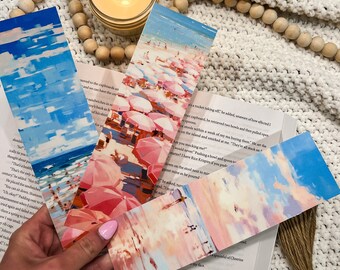 Puede incluir: Un conjunto de cuatro marcapáginas coloridos que representan una escena de playa con sombrillas rosas y un cielo azul. Los marcapáginas están hechos de papel y tienen un aspecto pintado.
