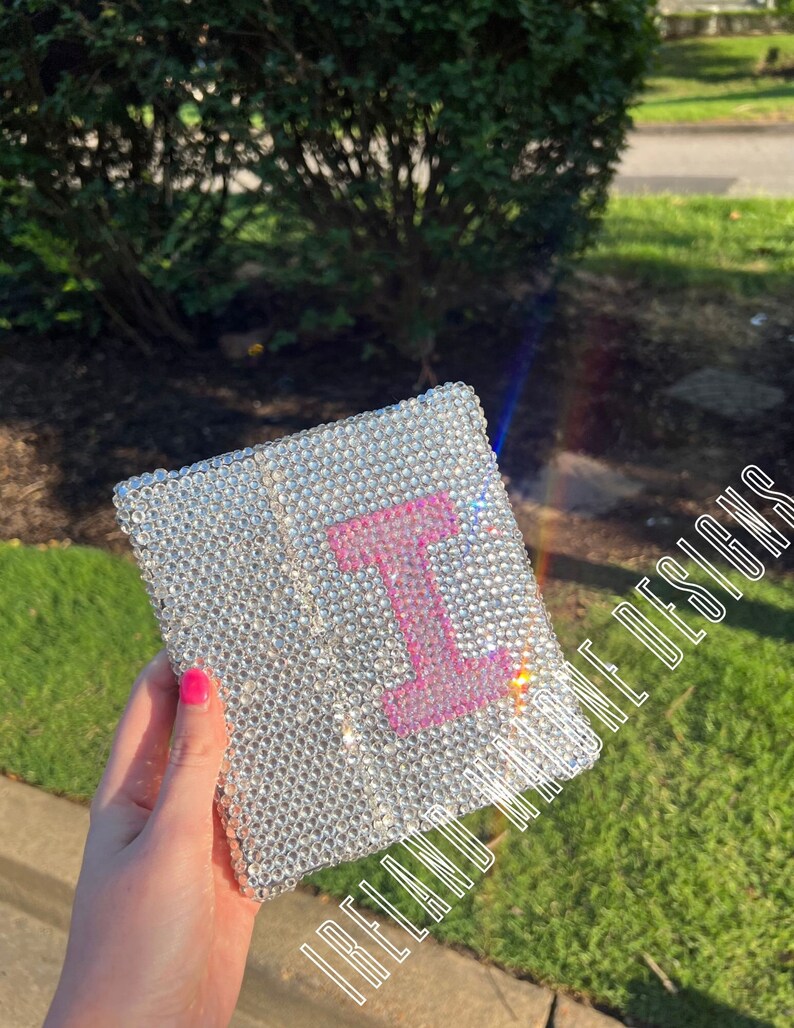 May include: A small, silver, sparkly notebook with a pink letter "I" on the front. The notebook is covered in rhinestones.