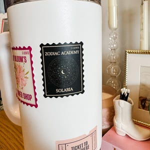 Puede incluir: Taza de viaje blanca con tres pegatinas. Las pegatinas son un dise&ntilde;o floral rosa con el texto "5 CENTS BLOOM'S WER SHOP", una pegatina en blanco y negro con el texto "5 CENTS ZODIAC ACADEMY SOLARIA", y una pegatina rosa con el texto "READERS ONLY TICKET TO ANYWHERE READERS ONLY".