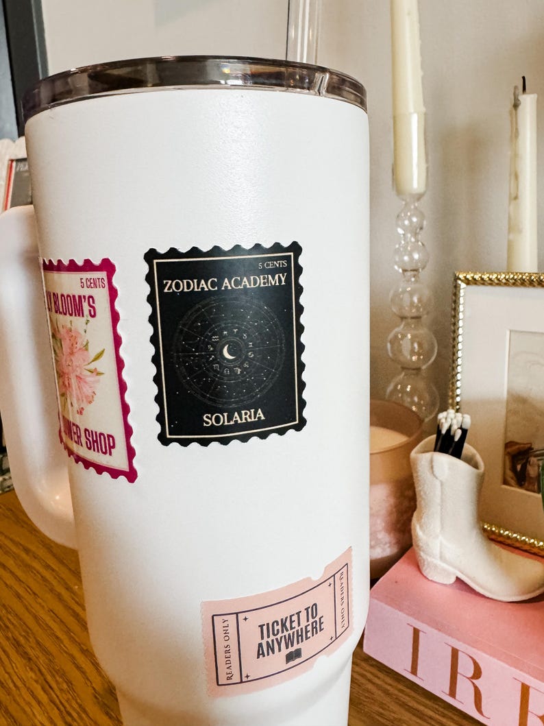 Puede incluir: Taza de viaje blanca con tres pegatinas. Las pegatinas son un dise&ntilde;o floral rosa con el texto "5 CENTS BLOOM'S WER SHOP", una pegatina en blanco y negro con el texto "5 CENTS ZODIAC ACADEMY SOLARIA", y una pegatina rosa con el texto "READERS ONLY TICKET TO ANYWHERE READERS ONLY".