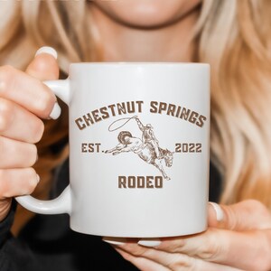 May include: White ceramic mug with a brown graphic of a cowboy riding a bucking horse. The text "Chestnut Springs Est. 2022 Rodeo" is printed on the mug.
