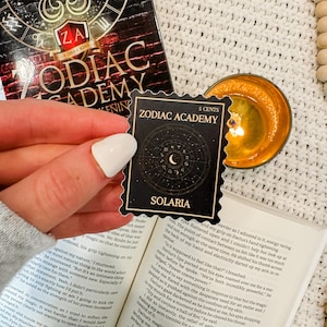 Puede incluir: Una pegatina negra y dorada con el texto "Zodiac Academy" y "Solaria" en letras blancas. La pegatina es un sello de 5 c&eacute;ntimos con un dise&ntilde;o de luna creciente y estrellas.