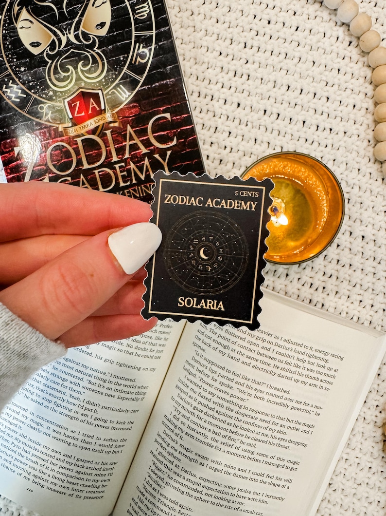 Puede incluir: Una pegatina negra y dorada con el texto "Zodiac Academy" y "Solaria" en letras blancas. La pegatina es un sello de 5 c&eacute;ntimos con un dise&ntilde;o de luna creciente y estrellas.