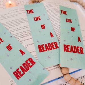 Puede incluir: Marcadores de libros azules claros con el texto "THE LIFE OF A READER" en rojo. Los marcadores est&aacute;n colocados sobre un libro abierto, con una vela y cuentas de madera en el fondo.