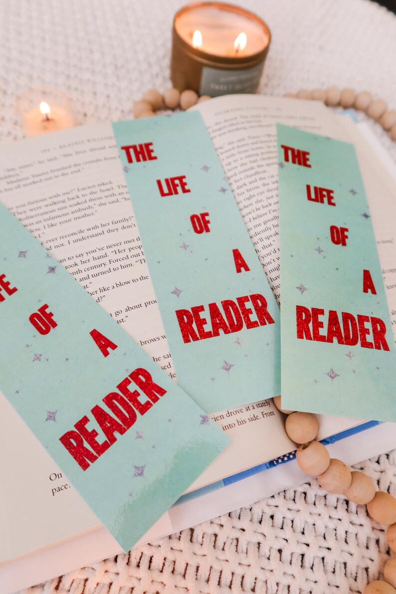 Puede incluir: Marcadores de libros azules claros con el texto "THE LIFE OF A READER" en rojo. Los marcadores est&aacute;n colocados sobre un libro abierto, con una vela y cuentas de madera en el fondo.