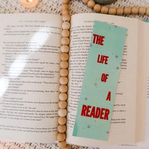 Puede incluir: Un libro abierto con un marcap&aacute;ginas turquesa que dice "The Life of a Reader" en texto rojo. Una guirnalda de cuentas de madera y velas encendidas rodean el libro.