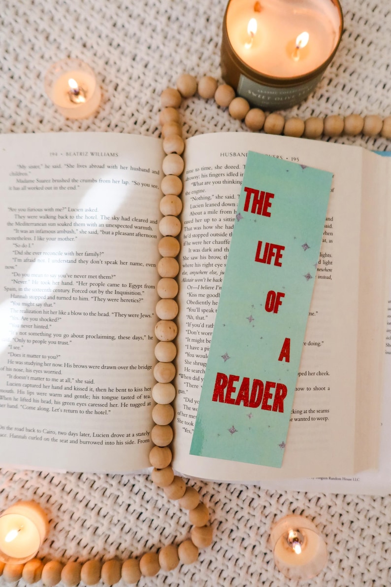 Puede incluir: Un libro abierto con un marcap&aacute;ginas turquesa que dice "The Life of a Reader" en texto rojo. Una guirnalda de cuentas de madera y velas encendidas rodean el libro.