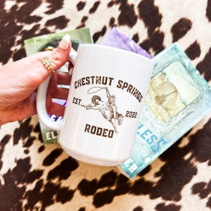 May include: White ceramic mug with a brown graphic of a cowboy riding a bucking bronco. The graphic includes the text "Chestnut Springs Rodeo Est. 2022".