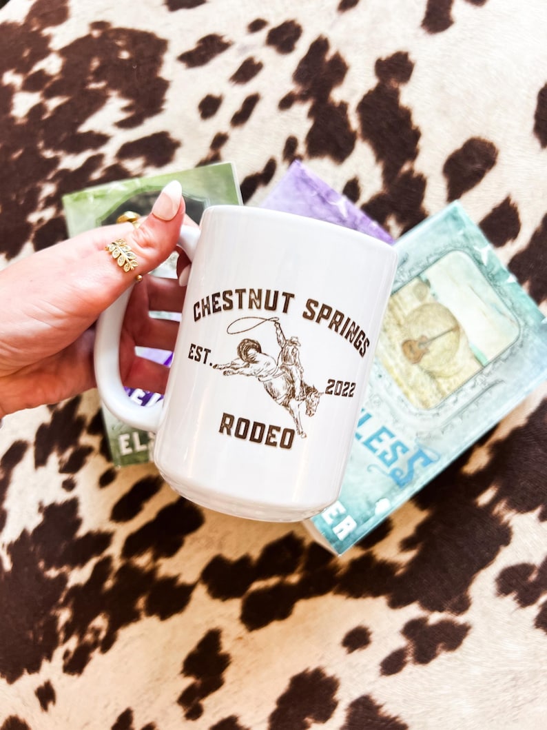 May include: White ceramic mug with a brown graphic of a cowboy riding a bucking bronco. The graphic includes the text "Chestnut Springs Rodeo Est. 2022".
