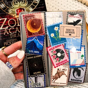 Puede incluir: Primer plano de una funda de tel&eacute;fono decorada con pegatinas coloridas. Las pegatinas presentan varios dise&ntilde;os, incluyendo una luna creciente, un horizonte urbano, un tocadiscos, una rosa salvaje y un billete que dice "Ticket to Anywhere".