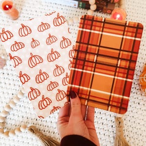 May include: Two rectangular notebooks. One features a white background with orange pumpkin outlines and polka dots. The other has an orange, red, and brown plaid pattern. The notebooks are held in a hand, with fall-themed decor in the background.