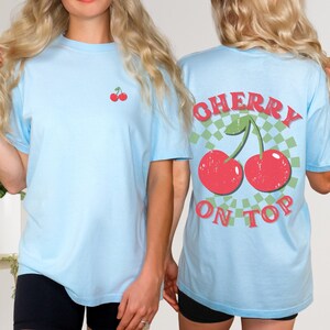 May include: Two light blue t-shirts with a cherry graphic. The front of one shirt has a small cherry graphic. The back of the other shirt has a larger cherry graphic with the text "CHERRY ON TOP".