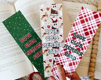 May include: Three Christmas-themed bookmarks with the text "BOOK CLUB MEMBER CARD". One is green with white stars, another has Santa and reindeer, and the third has a red plaid pattern. The bookmarks are next to an open book.