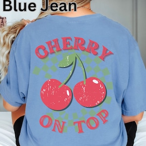 May include: A blue t-shirt with a graphic of two red cherries with green stems and leaves on a green and white checkered background. The text "CHERRY ON TOP" is printed in red letters on the shirt.