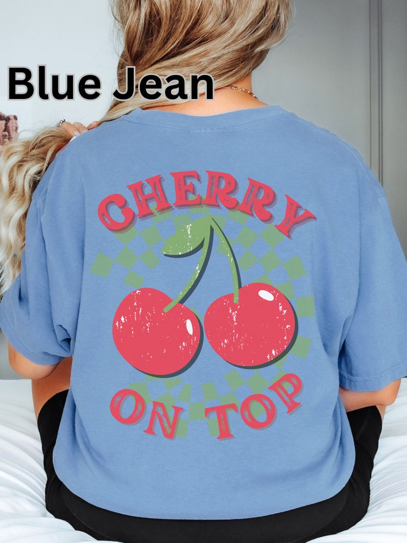 May include: A blue t-shirt with a graphic of two red cherries with green stems and leaves on a green and white checkered background. The text "CHERRY ON TOP" is printed in red letters on the shirt.