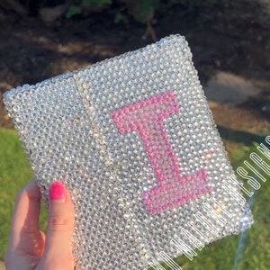 May include: A small, square, clear crystal-covered notebook with a pink letter "I" in the center. The notebook has a gold trim around the edges. The text "I Believe In Magic" is visible on the bottom right corner of the notebook.