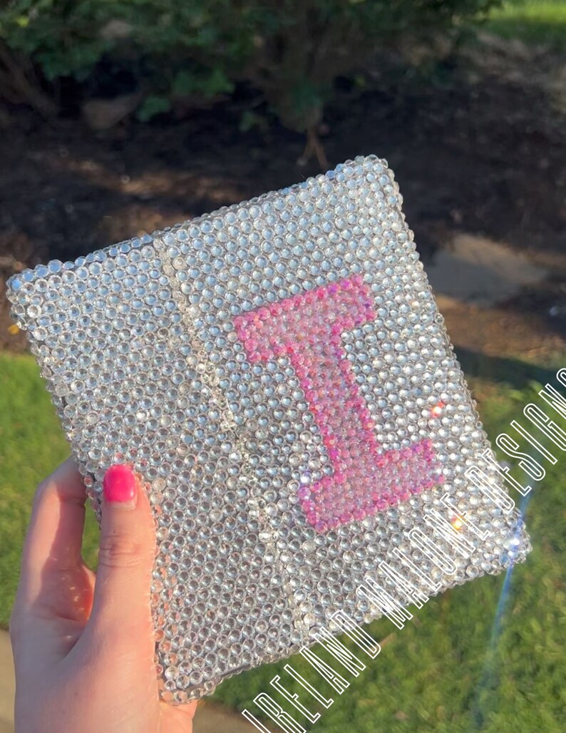 May include: A small, square, clear crystal-covered notebook with a pink letter "I" in the center. The notebook has a gold trim around the edges. The text "I Believe In Magic" is visible on the bottom right corner of the notebook.