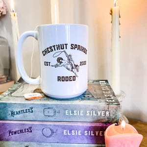 May include: White ceramic mug with a brown graphic of a cowboy riding a bucking bronco. The graphic includes the text "Chestnut Springs Est. 2022 Rodeo". The mug is sitting on a stack of three books with the title "Elsie Silver" on each book. There is a pink cowboy boot candle next to the mug.